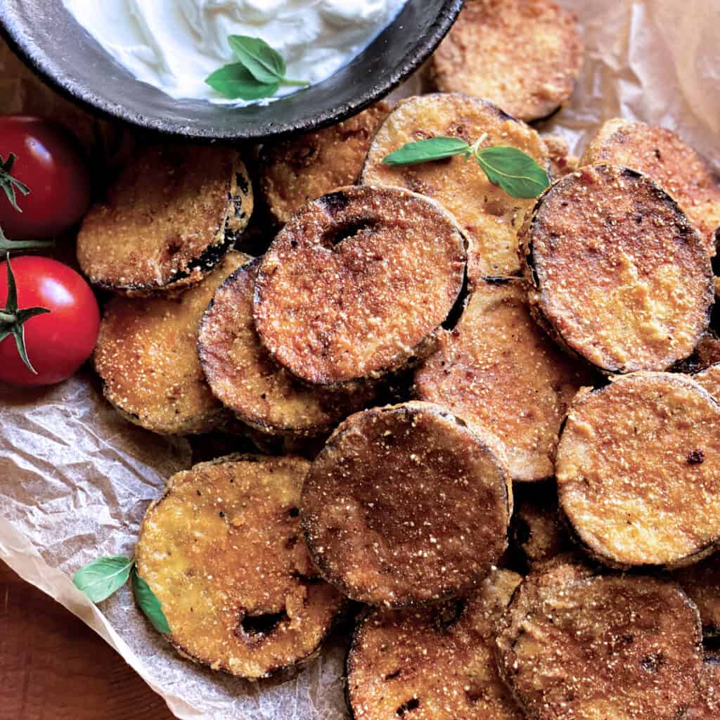 Fried eggplant slices on parchment paper, a couple of tomatoes and a bowl with yogurt dip.