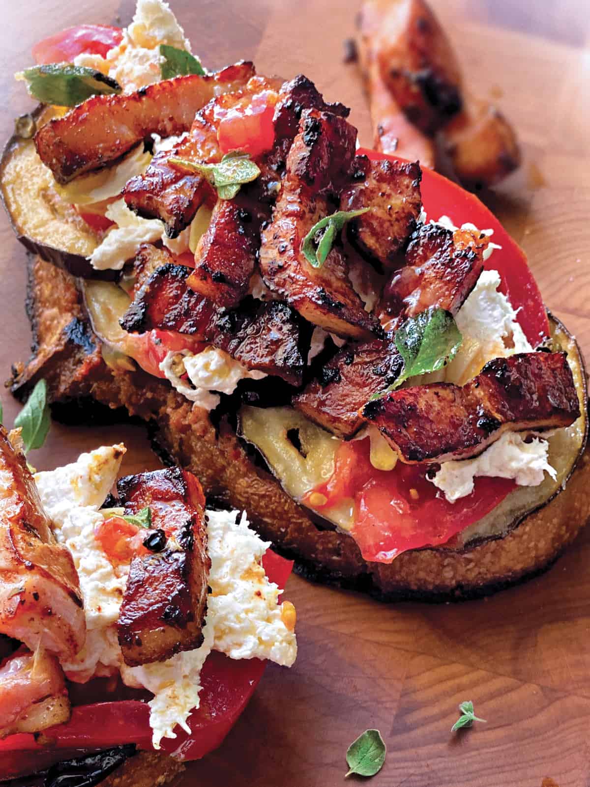 An open sandwich and part of another, both with with eggplant, tomatoes pancetta and mozzarella on a wooden surface.