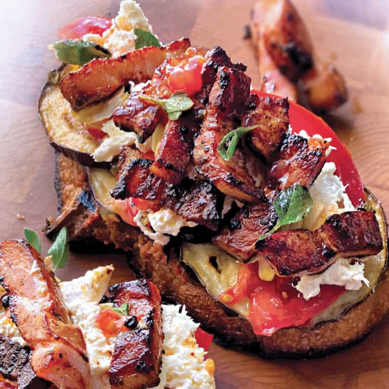 An open sandwich and part of another, both with with eggplant, tomatoes pancetta and mozzarella on a wooden surface.