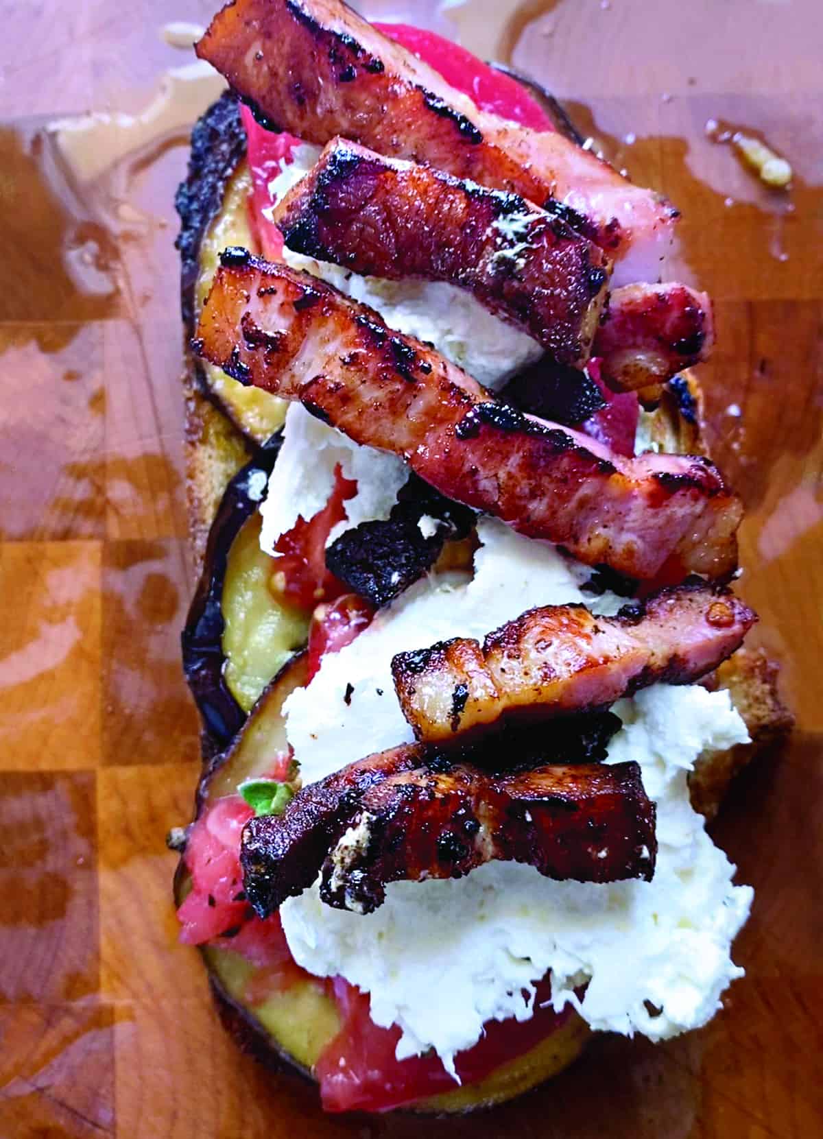 An open sandwich with with eggplant, tomatoes, pancetta and mozzarella on a wooden surface.