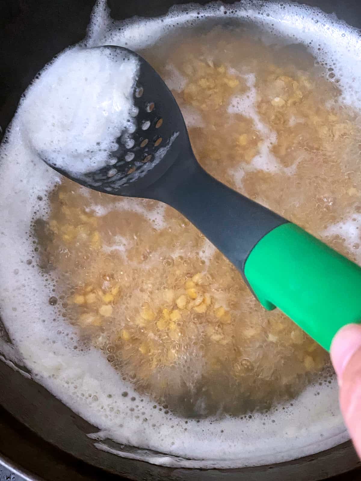 Yellow split peas boiling and a hand holding a spoon picks up foam.