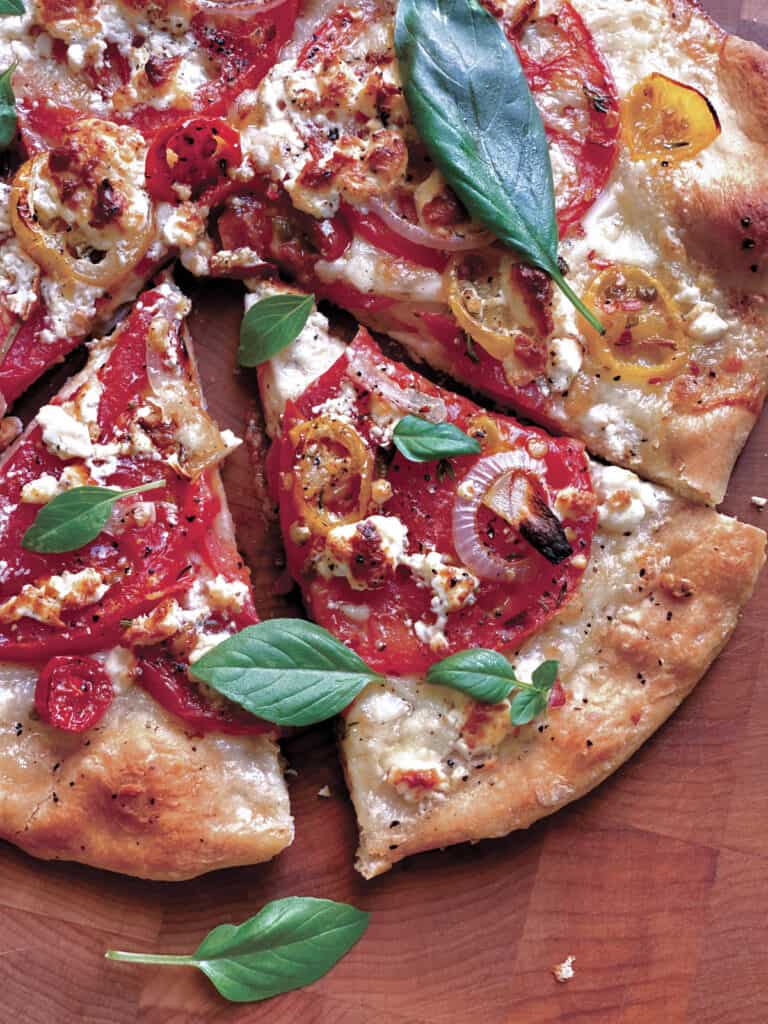 A feta pizza on a cutting board, two pieces cut and basil leaves scattered on top.