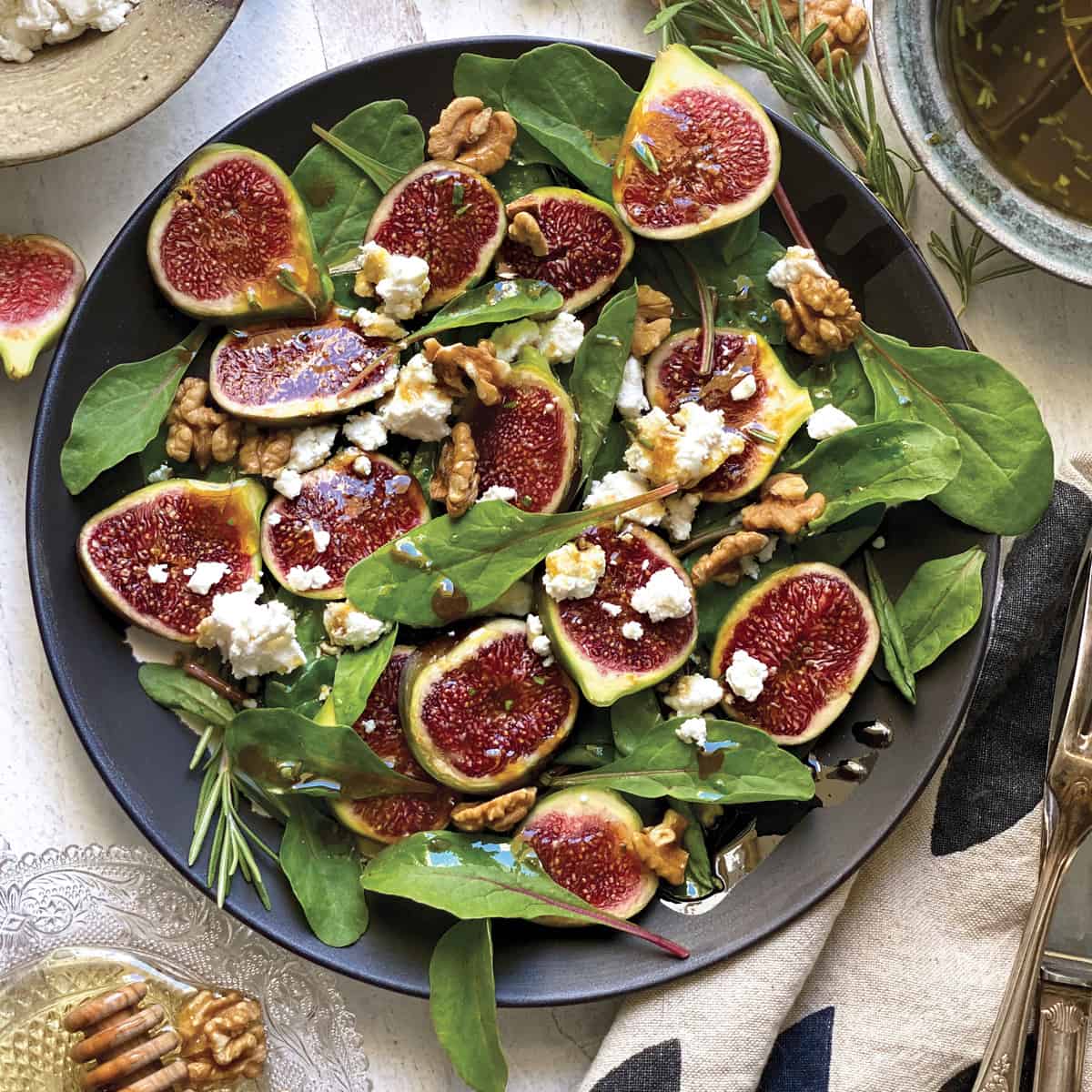 A plate with fig salad with rosemary vinaigrette surround by a cloth napkin, a bowl with dressing, utensils, figs, walnuts and rosemary on a white table.