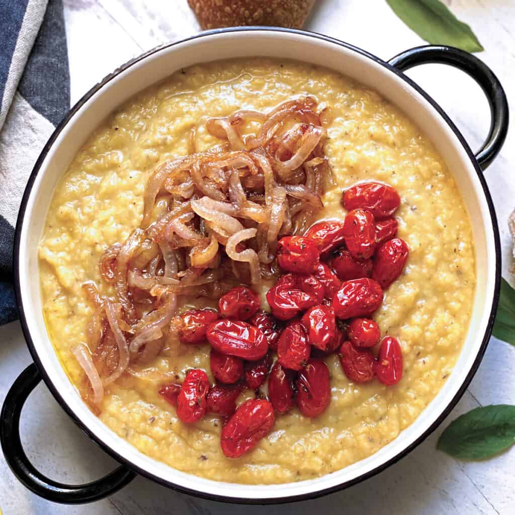 A serving plate with greek fave dip with tomatoes and onions, some pieces of bread, half a lemon and partial views of a pan with onions and a pan with tomatoes with a spoon.