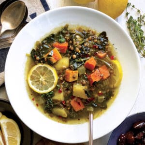A plate with lentil spinach soup and a spoon. Around it are a bowl with lemon wedges and a bowl with olives.