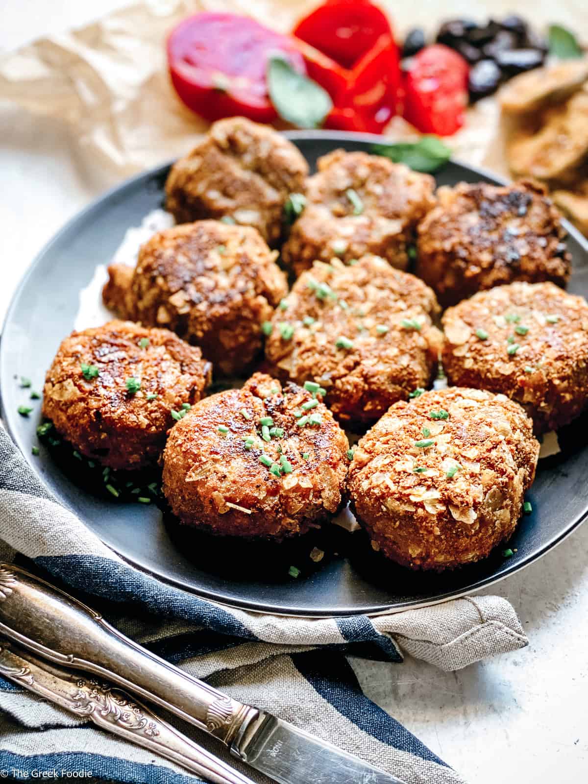 Nine crispy potato fritters on a dark plate garnished with chopped chives, with sliced tomatoes and fried eggplant in the background.