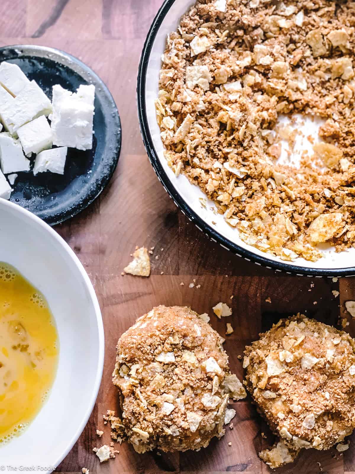 Overhead view of coated potato fritters on a wooden cutting board next to bowls of beaten egg, cubed feta cheese, and breadcrumb-chip coating mixture.