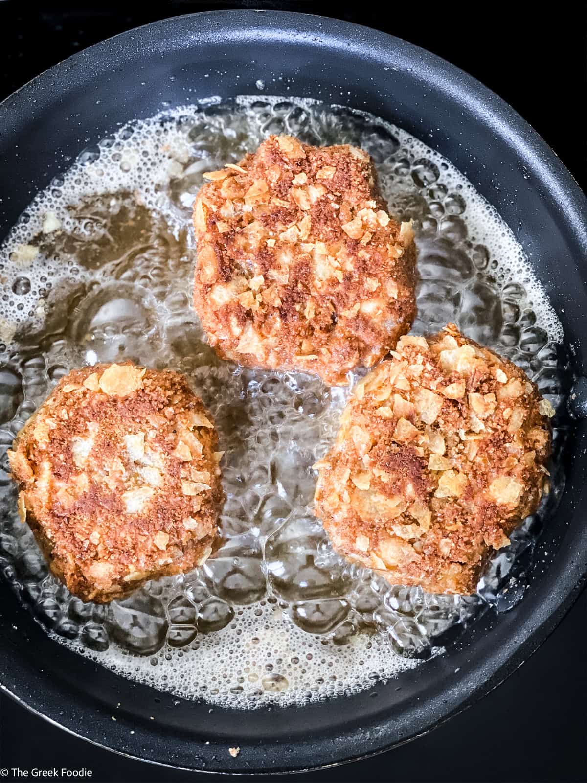 Three breaded potato fritters frying in hot oil in a dark skillet until golden brown.