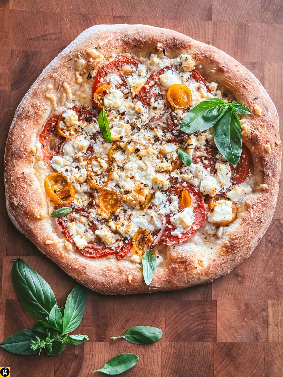 Whole feta pizza with tomatoes and basil on a butcher block, with fresh basil leaves beside it