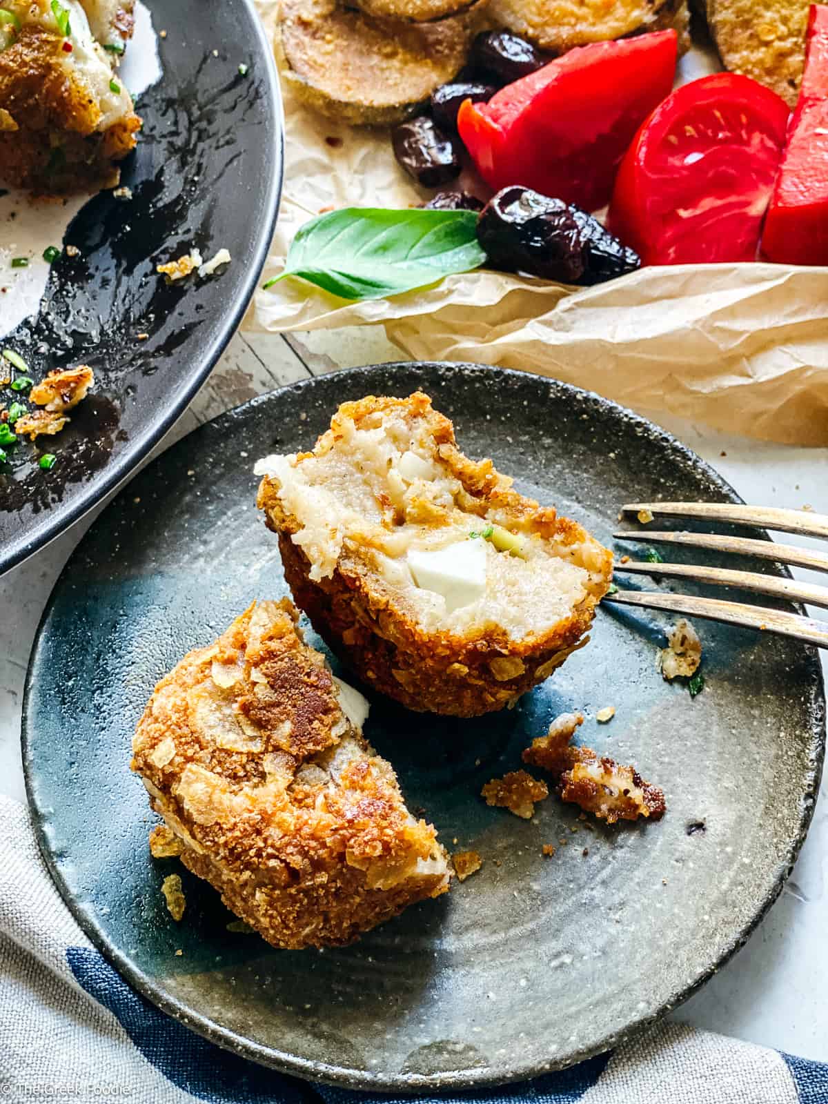 A crispy potato fritter cut in half on a small plate revealing the melted feta cheese center, with tomatoes, olives, and more fritters in the background.