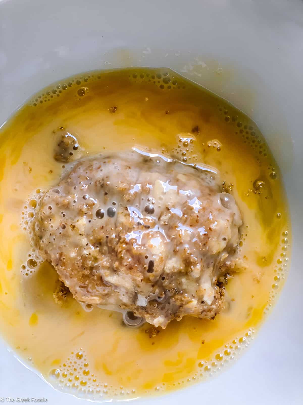 A breadcrumb-coated potato fritter being dipped into beaten egg for the second round of double-dip coating.
