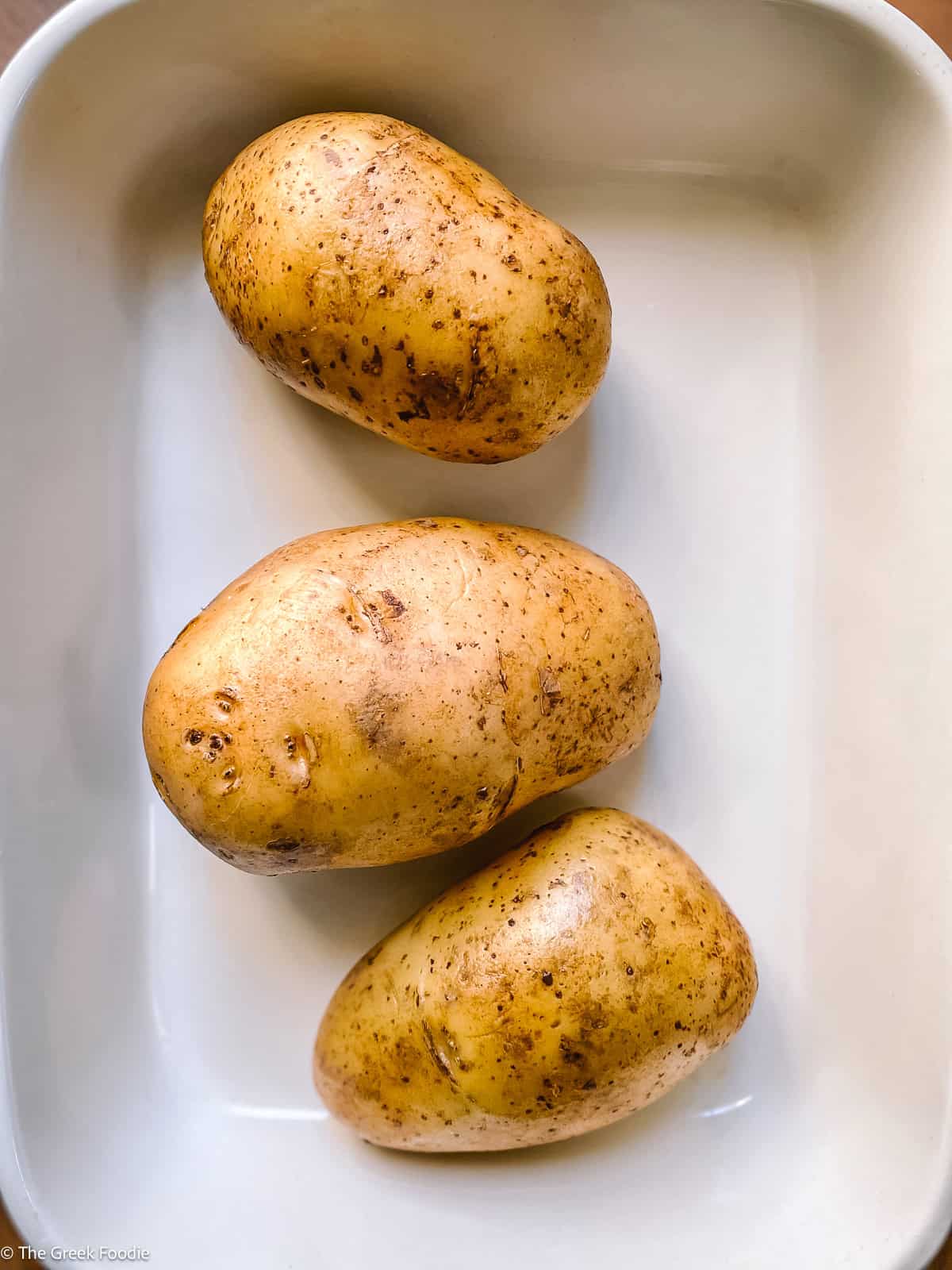 Three whole potatoes in a white baking dish, ready to be baked in the oven.