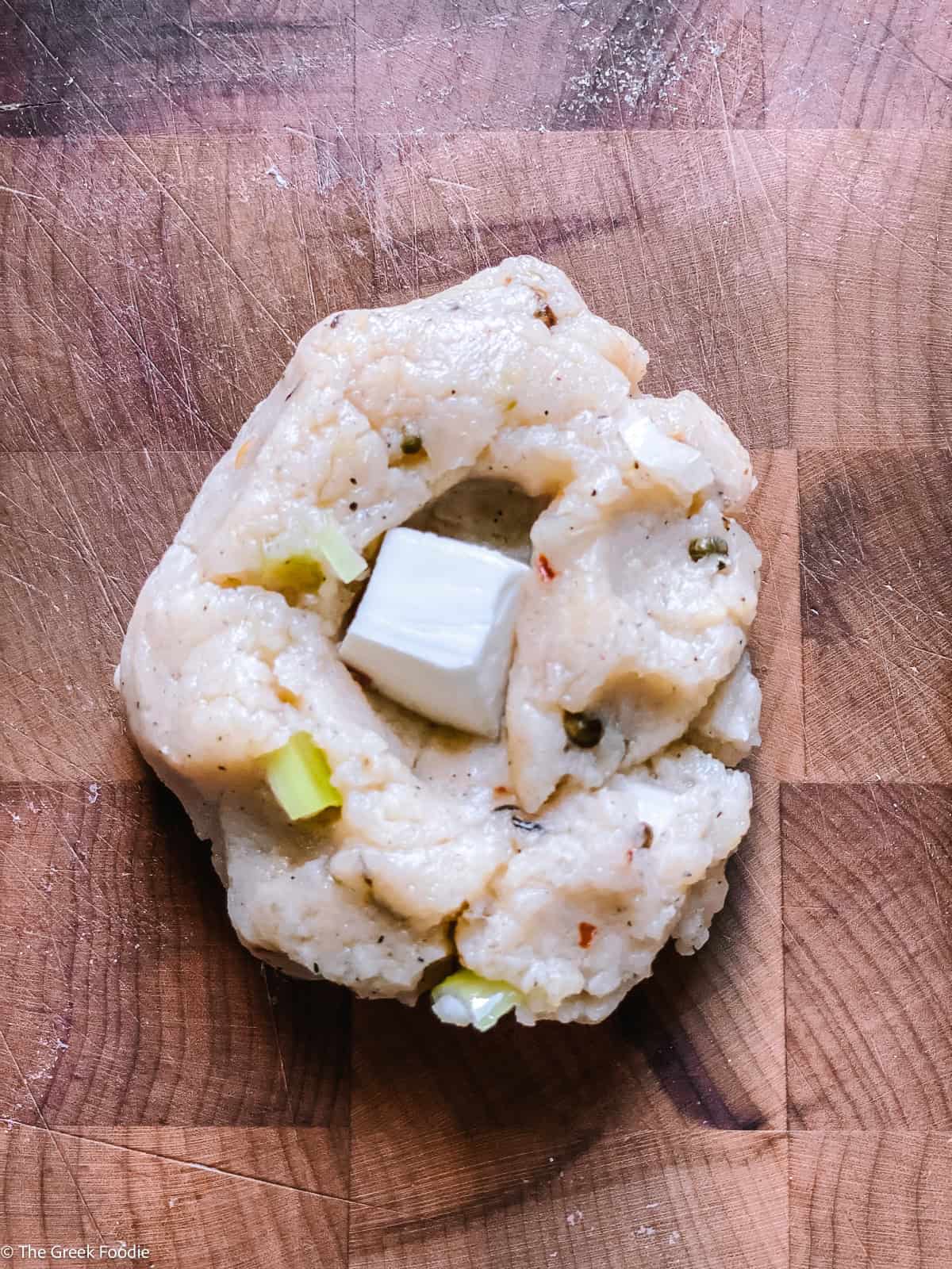 A flattened potato mixture with a cube of feta cheese pressed into the center on a wooden cutting board, ready to be sealed.