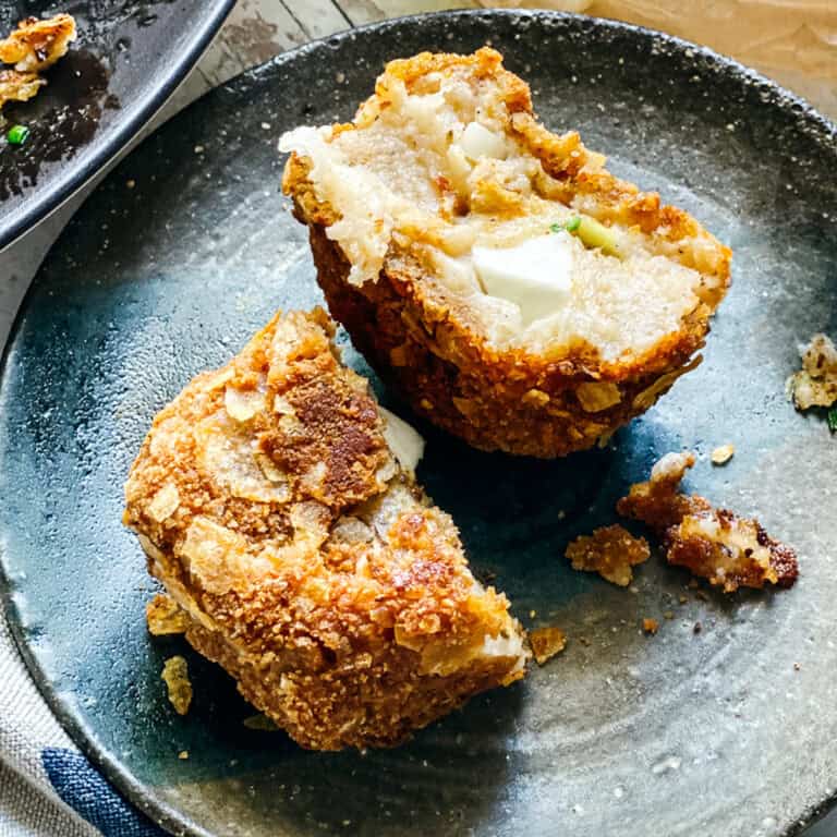 A crispy potato fritter cut in half on a small plate revealing the melted feta cheese center, with tomatoes, olives, and more fritters in the background.