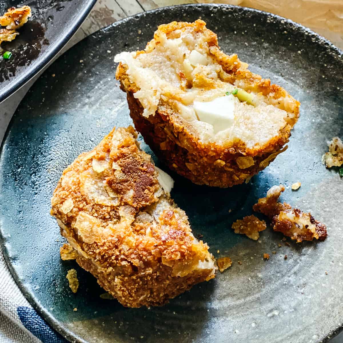 A crispy potato fritter cut in half on a small plate revealing the melted feta cheese center, with tomatoes, olives, and more fritters in the background.