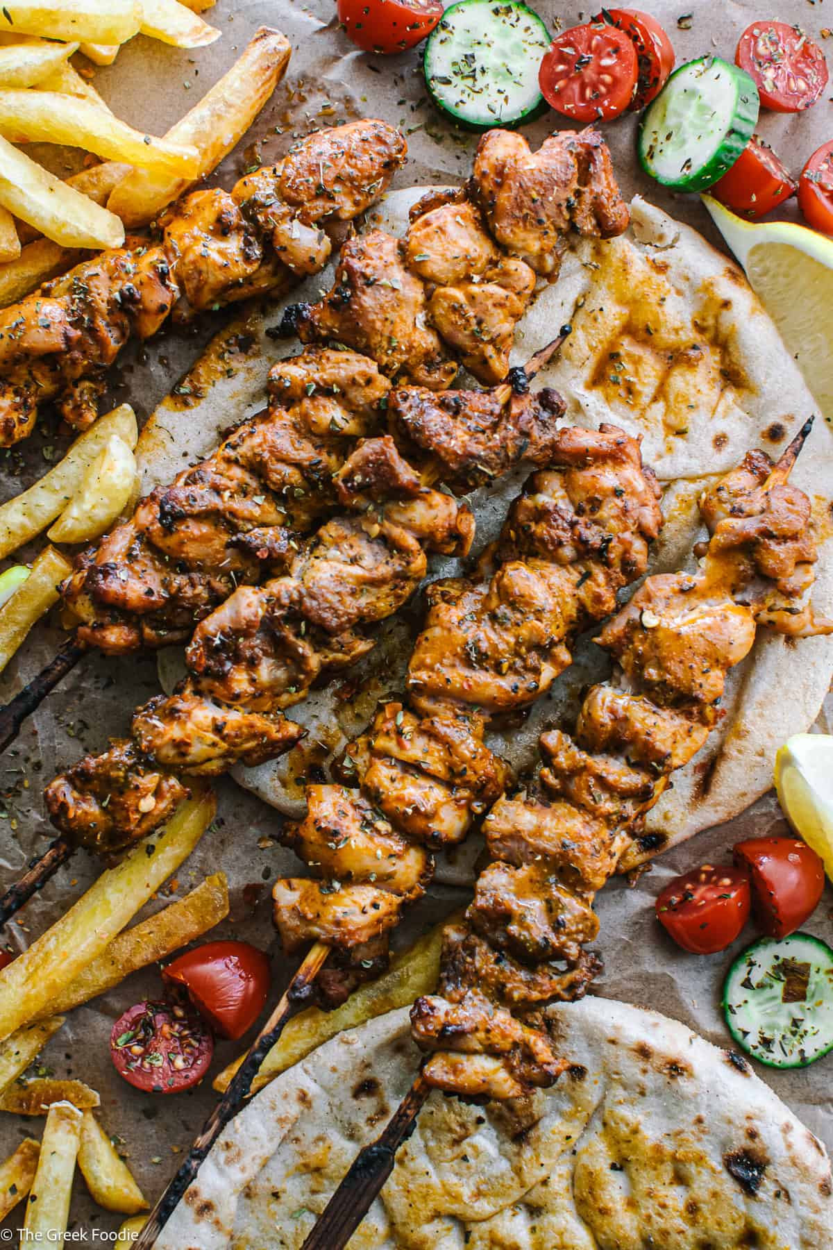 Chicken thigh skewers on pita, cherry tomatoes, sliced cucumber, fries and lemon wedges on parchment paper.