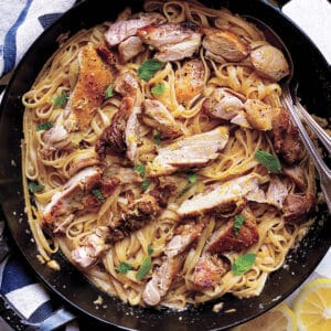 A skillet with lemon chicken pasta, utensils, herbs, a cloth napkin, and lemon halves.