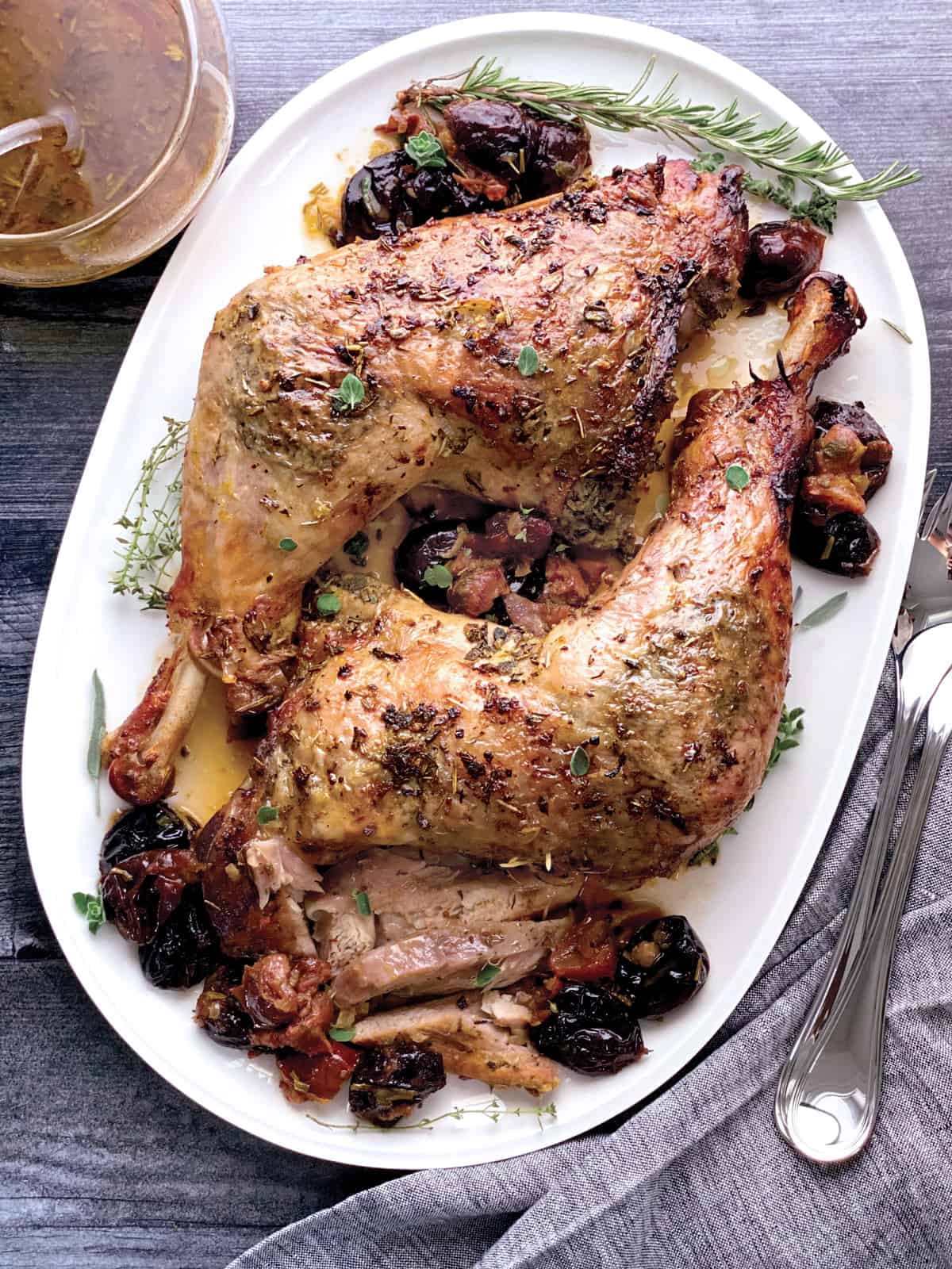 Two turkey thighs with drumsticks on a platter with herbs and dry fruit, and gravy. Serving utensils are on one side and a bowl with gravy on the other.