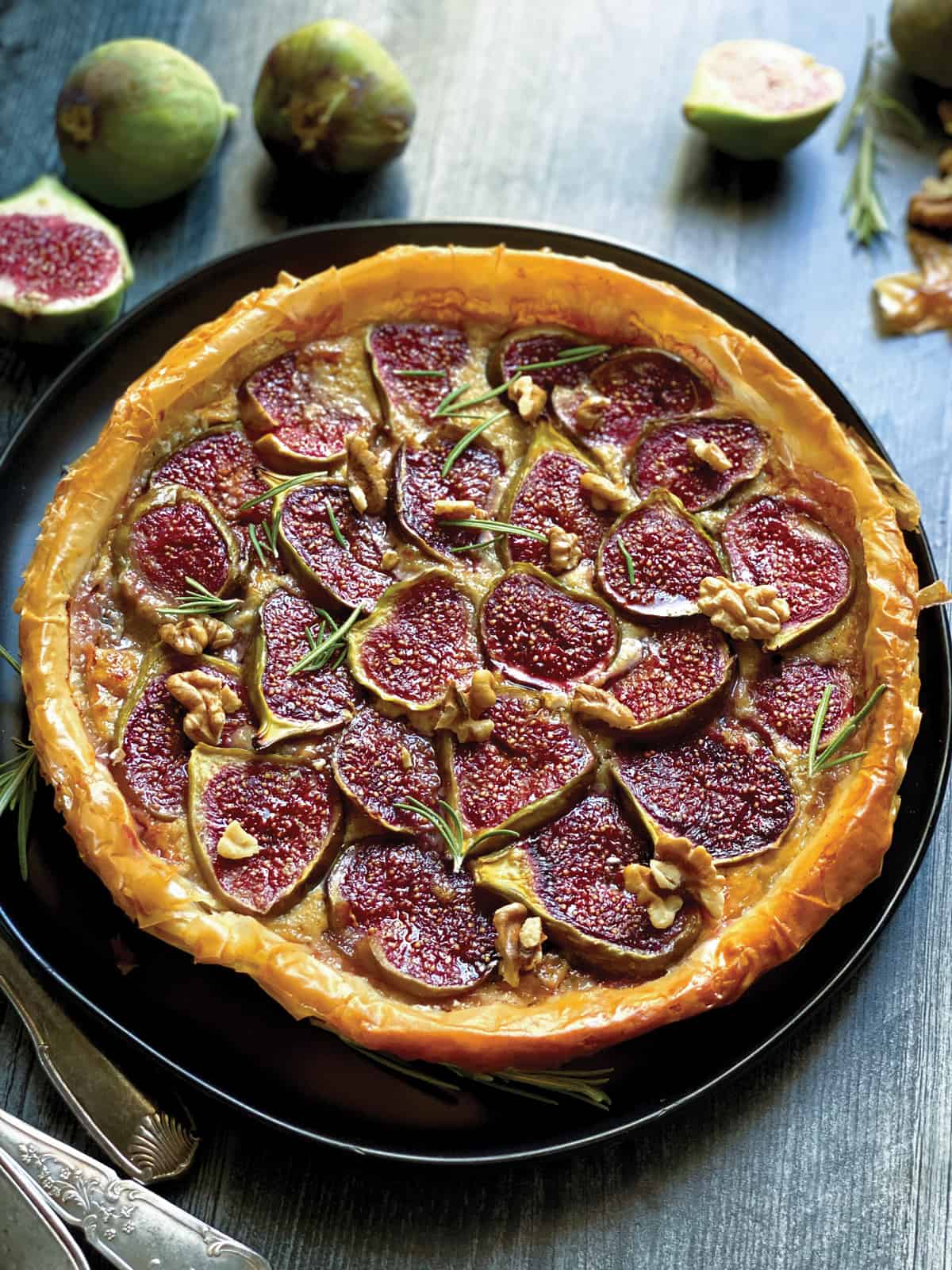 A phyllo tart with figs, utensils and  fig halves on a dark table.