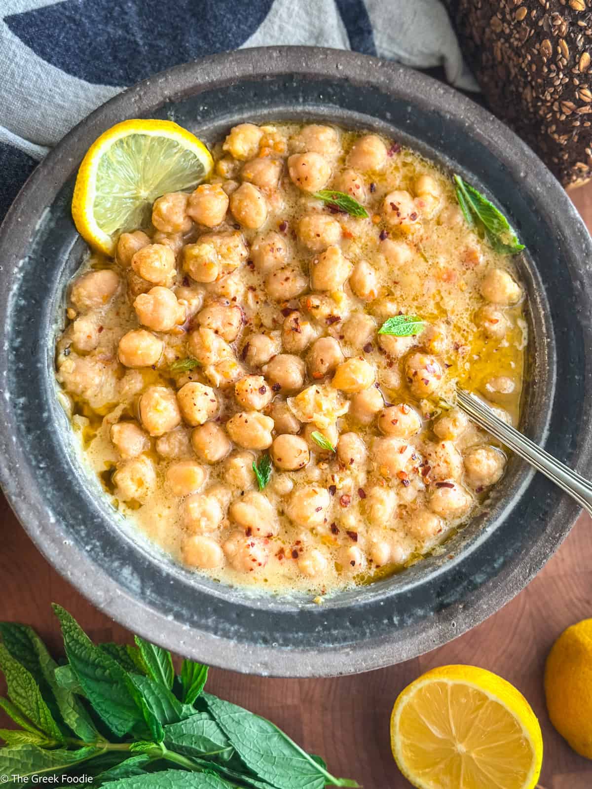 A bowl with chickpea soup, and a lemon slice, a cloth napkin, lemons and herbs on a table.
