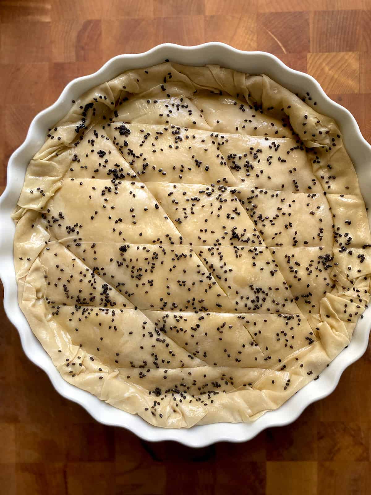 A greek pie with phyllo and sesame seeds on a butcher block ready for the oven.