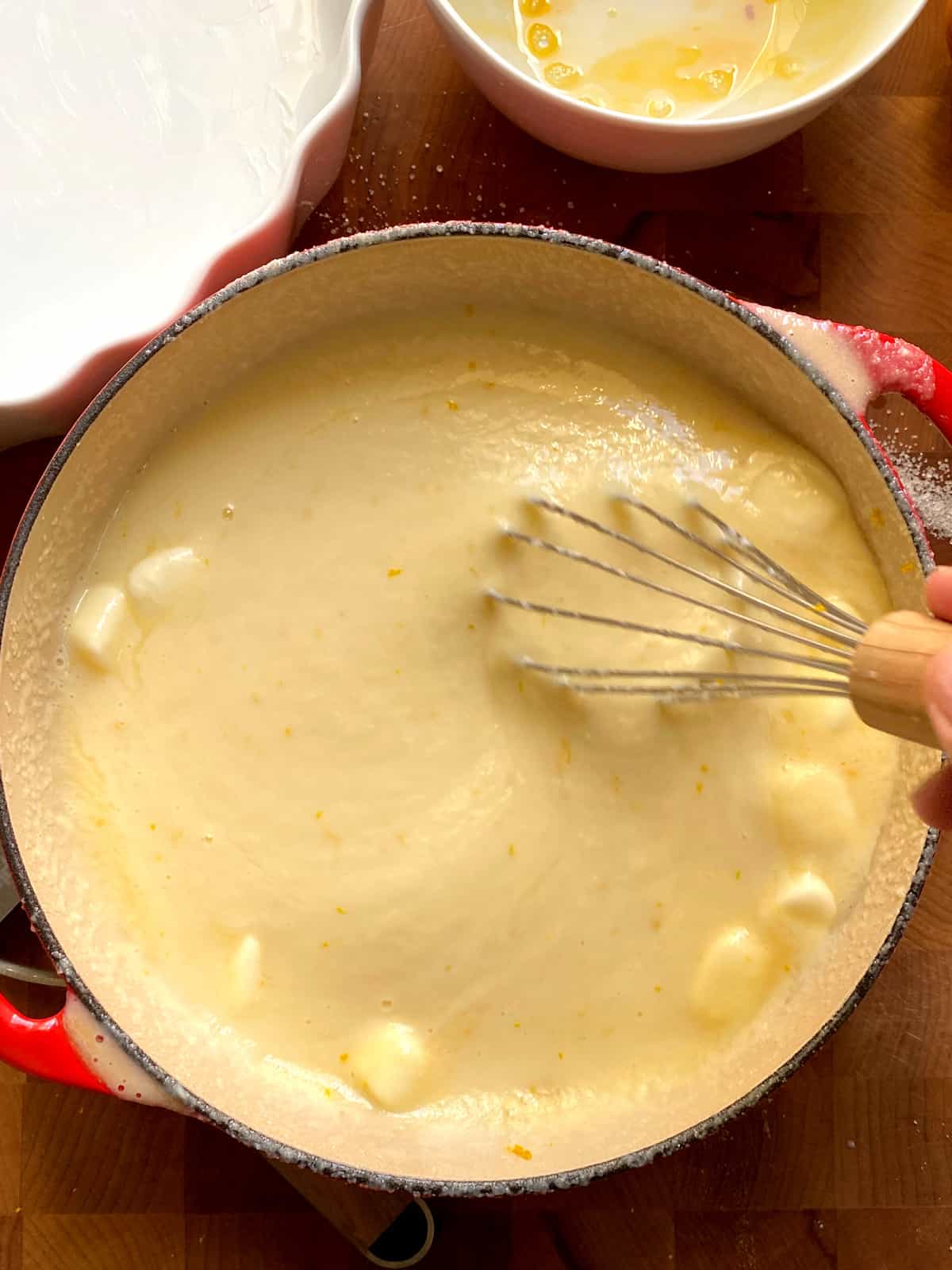 A stock pot on wood, a hand whisking custard with butter, an empty bowl above and partial view of a white tart pan on the left.