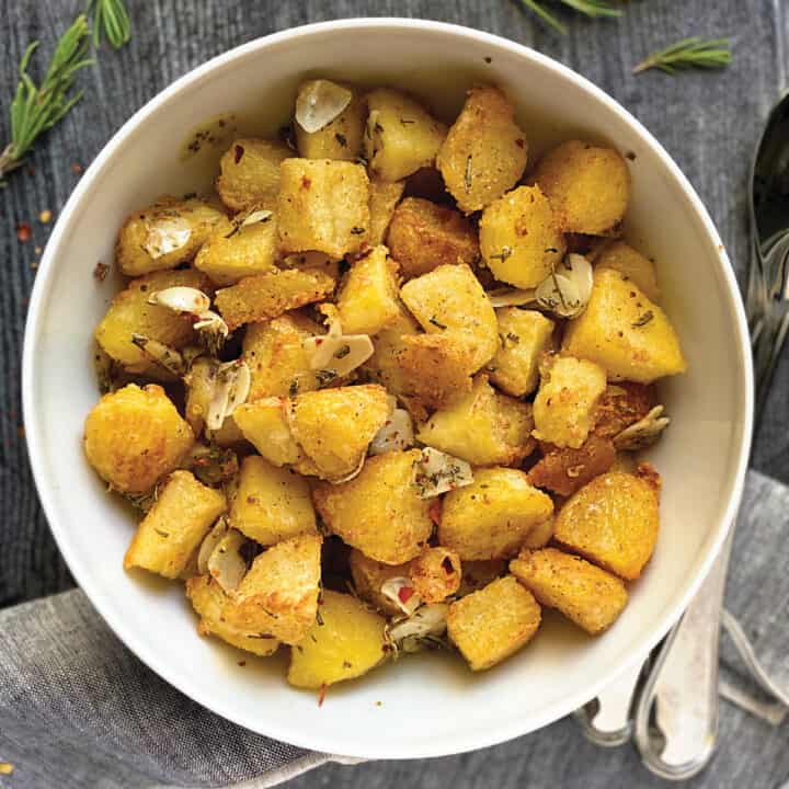 A white bowl with rosemary potatoes with garlic and parmesan on a grey wooden table with serving utensils and a cloth napkin.