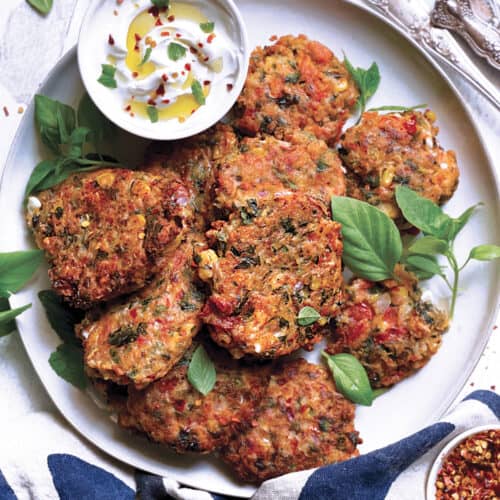 A plate with tomato corn fritters, utensils on a cloth napkin and a bowl with yogurt sauce at the back.