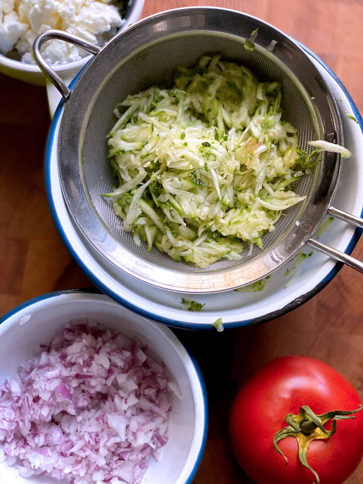 Grated zucchini in a strainer over a bowl, next to a bowl with onion, and a tomato.