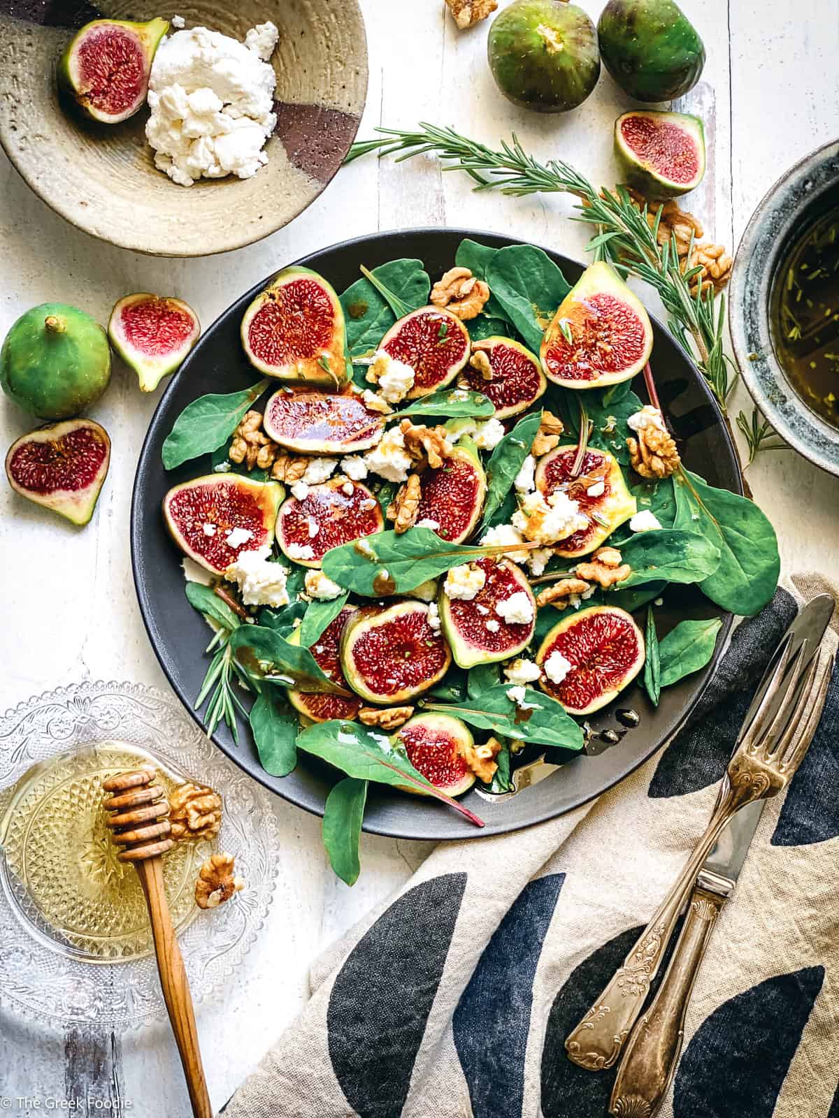 Fresh fig salad with halved figs, crumbled mizythra and walnuts on a dark plate, with rosemary vinaigrette and a honey dipper alongside.