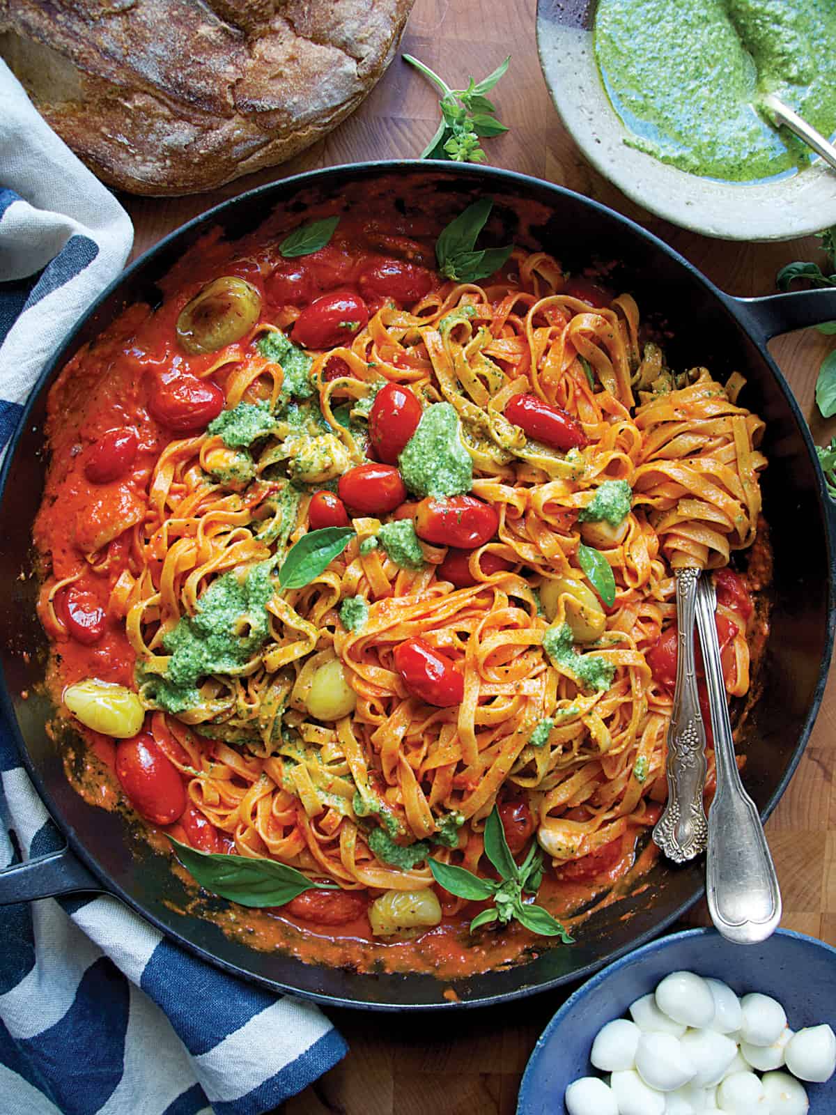 A cast iron pan with cherry tomato pasta sauce, serving utensils, partial view of a bread loaf and a bowl with pesto.