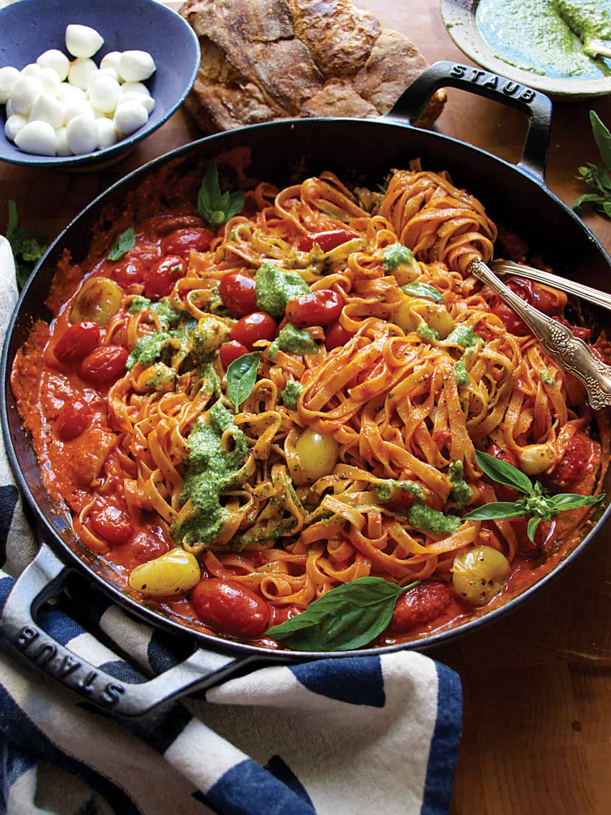 A cast iron pan with cherry tomato pasta sauce with pesto, serving utensils, partial view of a bread loaf and a bowl with pesto.