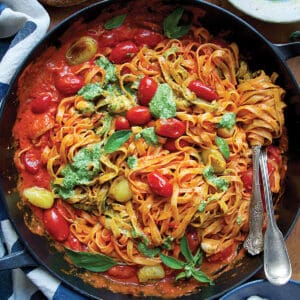 A cast iron pan with cherry tomato pasta sauce with pesto, serving utensils, partial view of a bread loaf and a bowl with pesto.