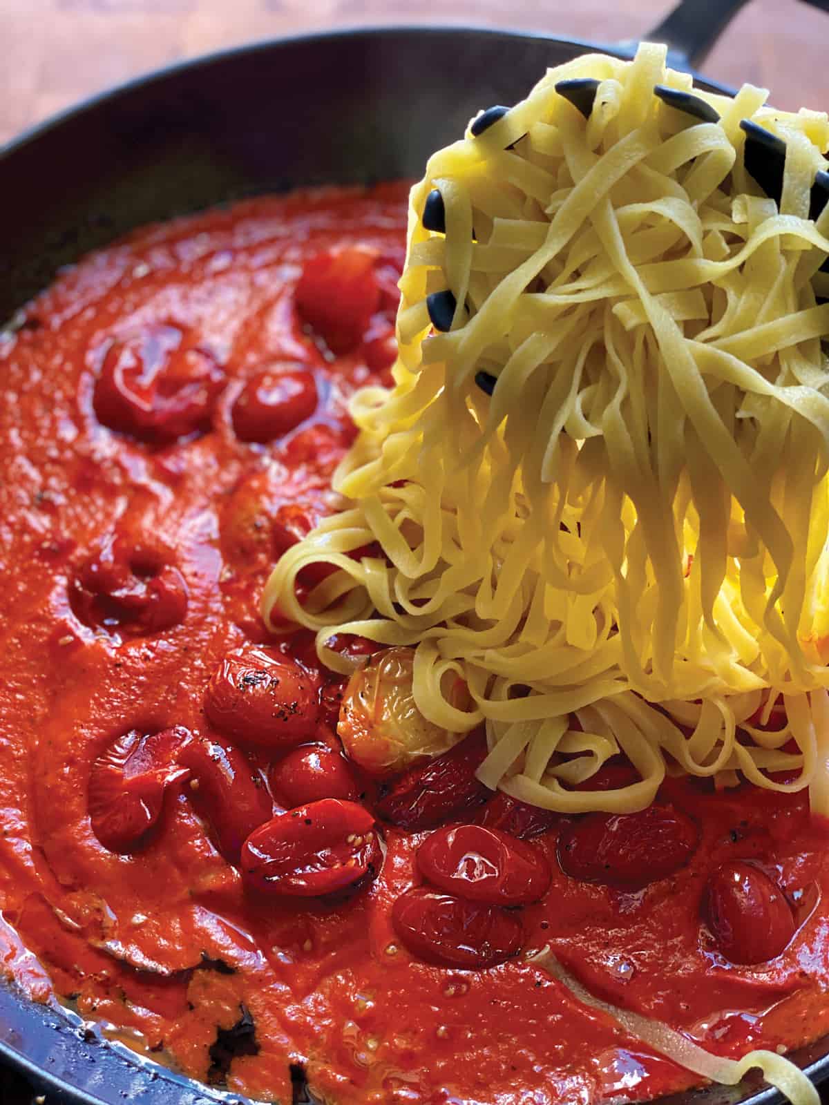 Pasta is being mixed with creamy tomato sauce mixed with roasted cherry tomatoes in a cast iron pan.