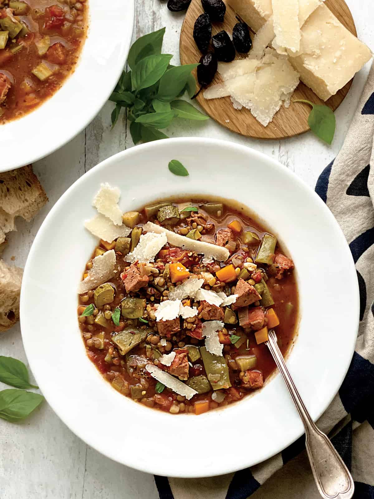 A bowl with lentil sausage soup, partial view of another bowl, some bread, and a tray with olives and parmesan cheese.