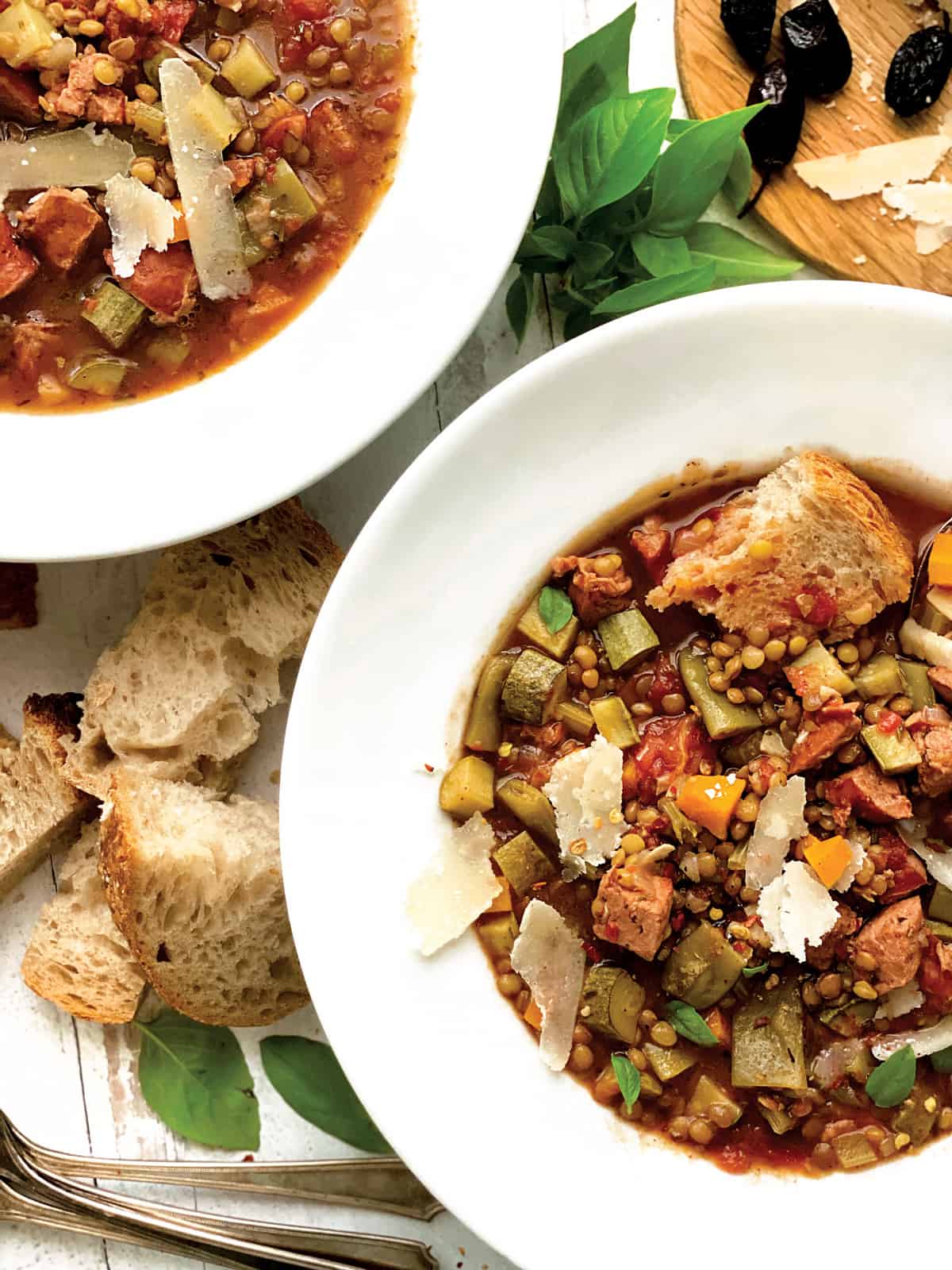 A bowl with lentil sausage soup, partial view of another bowl, some bread, and a tray with olives and parmesan cheese.