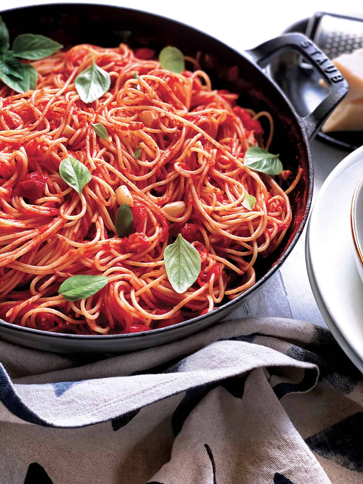 A large deep skillet with tomato garlic pasta and fresh basil. Partial view of parmesan and a grater and pasta bowls.