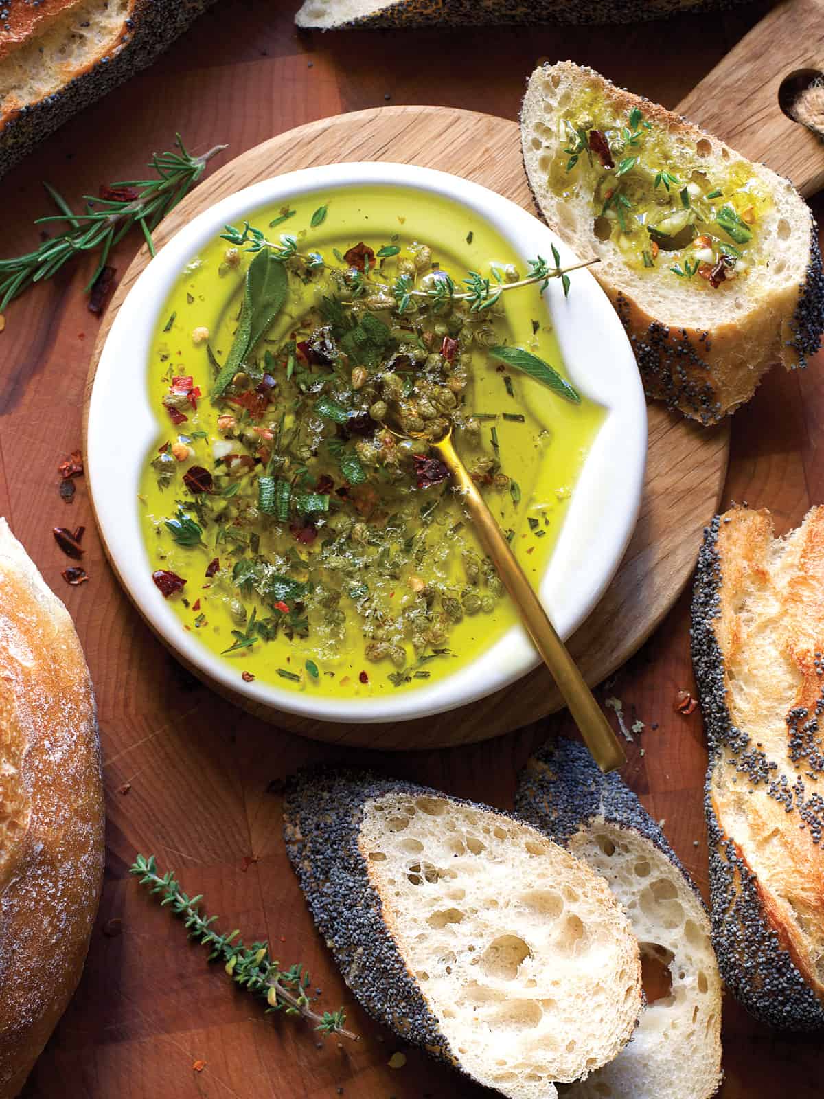 A small plate with olive oil and herbs, a gold spoon on a butcher block surrounded by pieces of bread.
