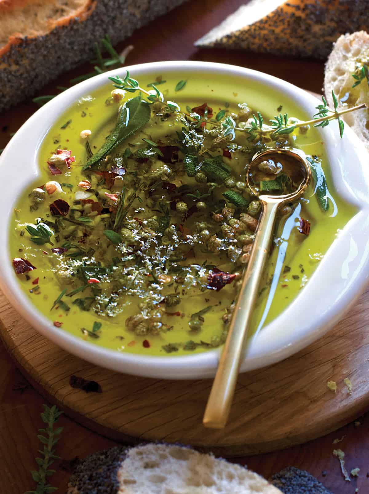 A small plate with olive oil and herbs, a gold spoon on a butcher block surrounded by pieces of bread.