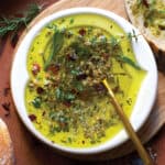 A small plate with olive oil and herbs, a gold spoon on a butcher block surrounded by pieces of bread.