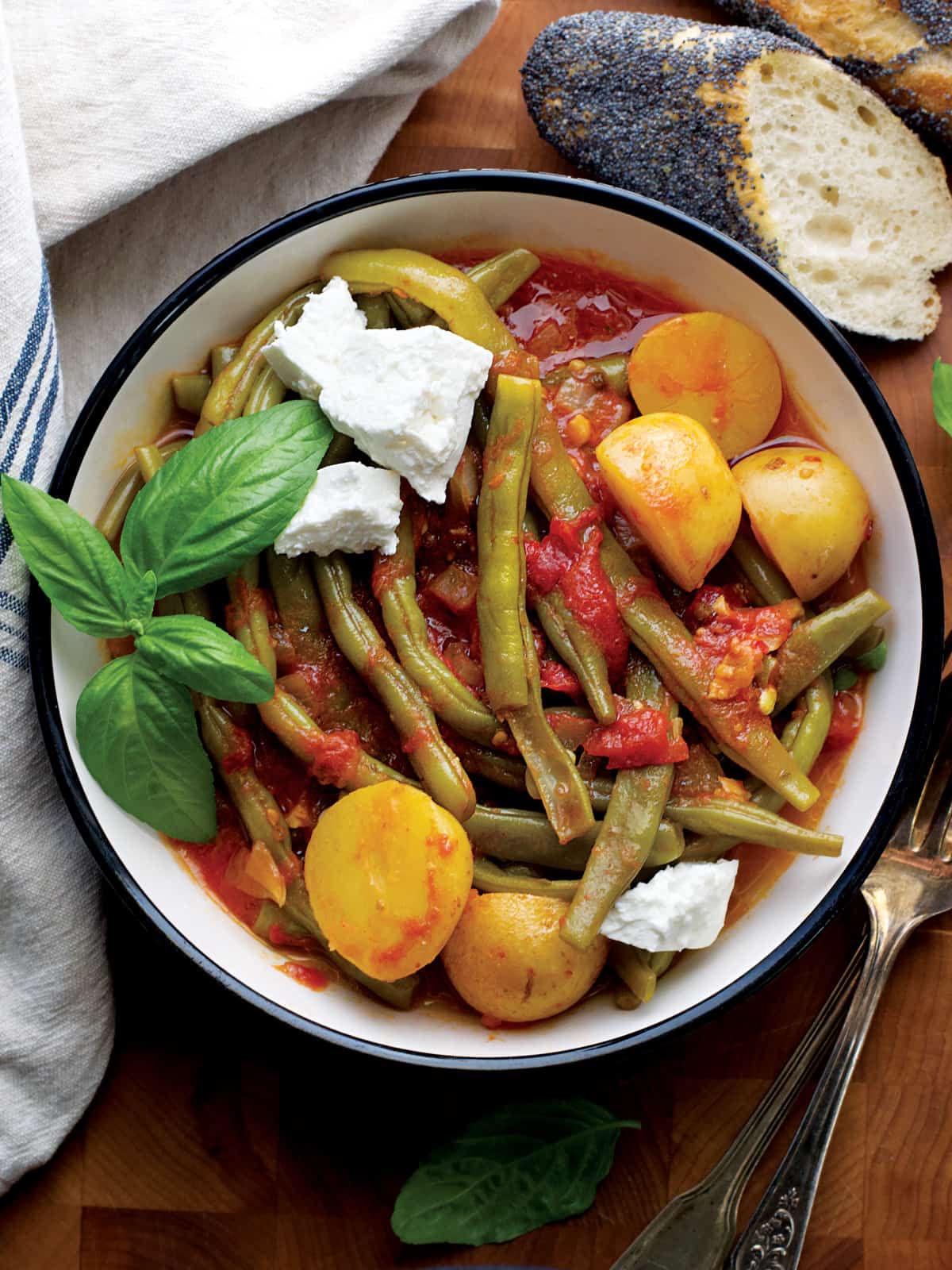 A plate with Greek green beans with potatoes in tomato sauce, some fresh basil. On the table, a napkin, utensils and a piece of bread.