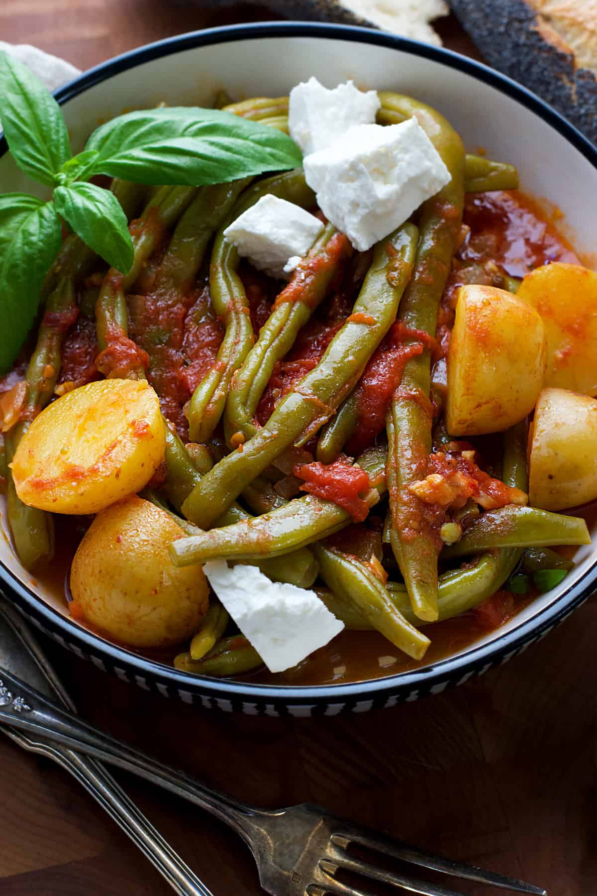 A plate with Greek green beans with potatoes in tomato sauce, a couple of feta cheese cubes and some fresh basil. On the table, utensils and a piece of bread.