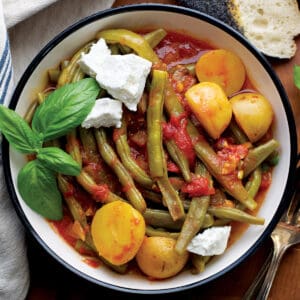 A plate with Greek green beans with potatoes in tomato sauce, some fresh basil. On the table, a napkin, utensils and a piece of bread.