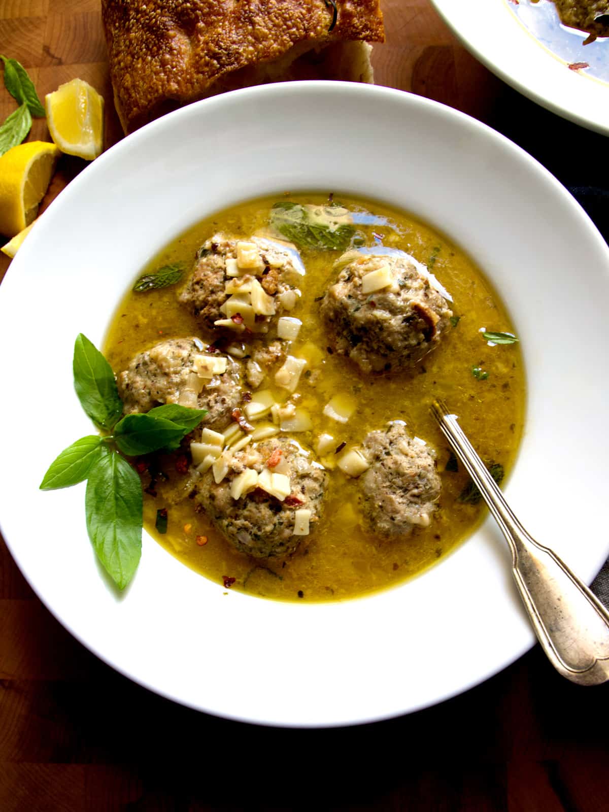 A bowl of meatball soup with some herbs and a spoon. Above it a loaf of bread and some lemon wedges.