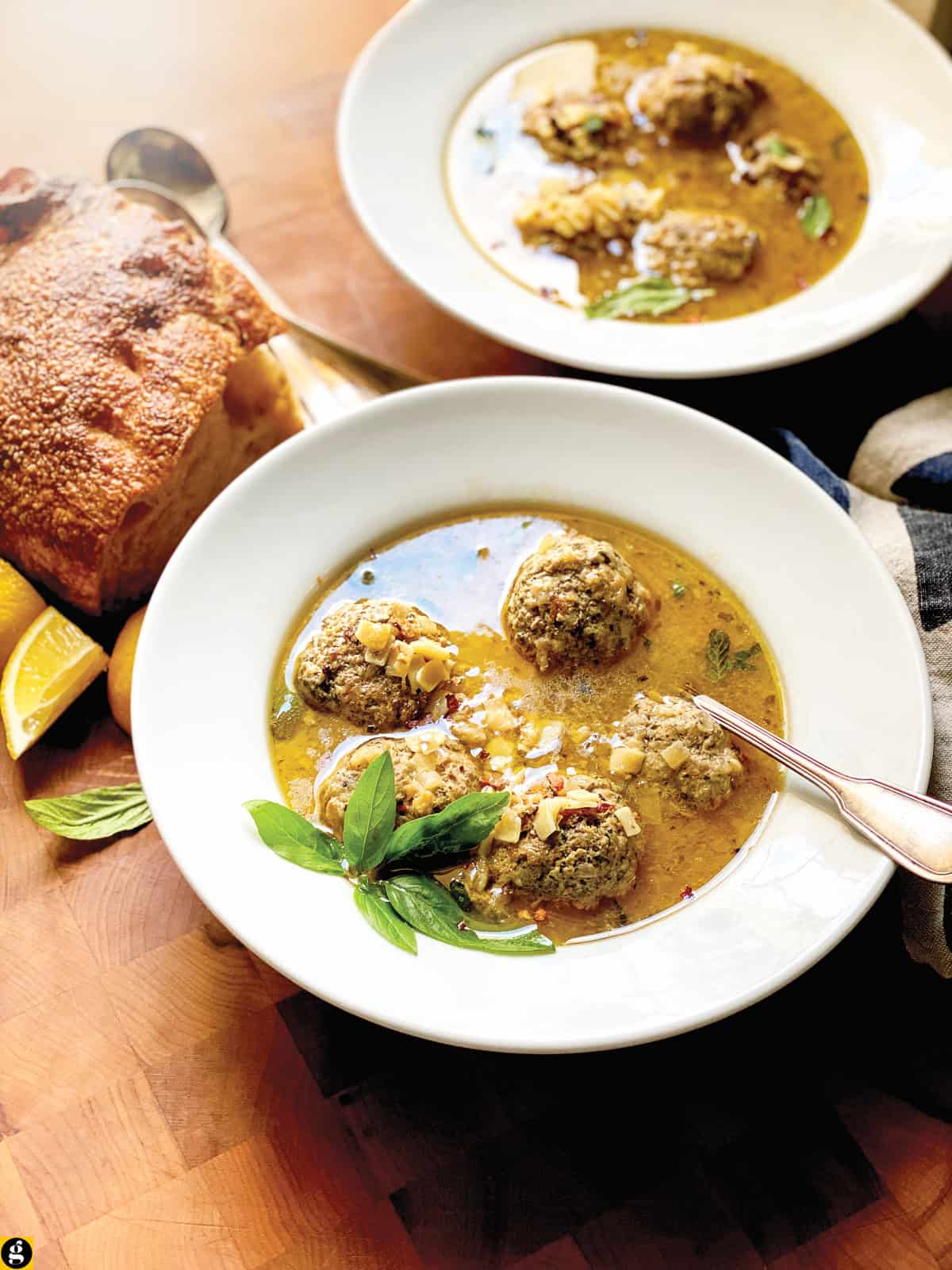 Side view of two soup bowls with meatball soup with tiny square pasta, basil, mint, and chili flakes in a lemony broth, with lemons and bread nearby.