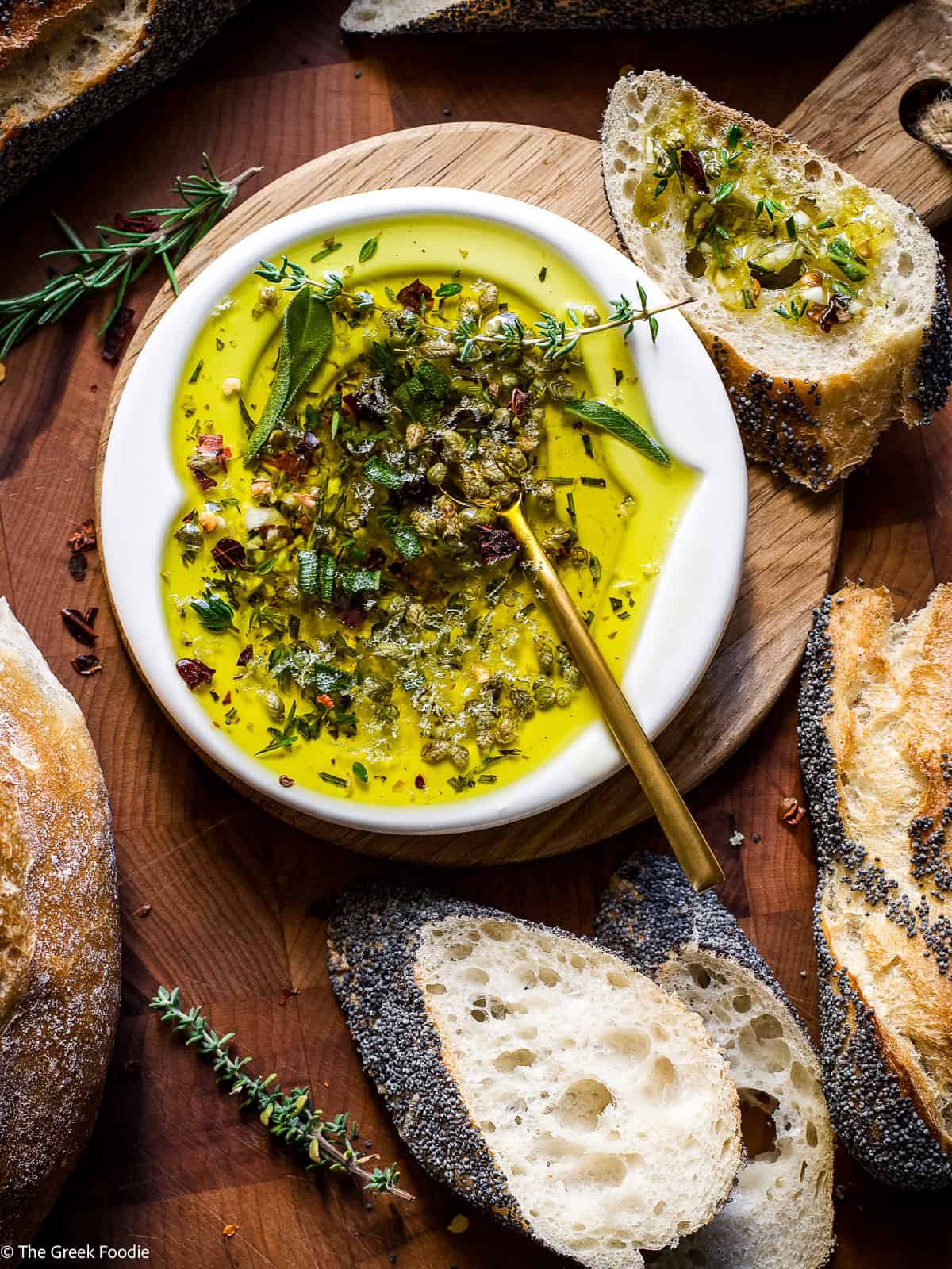 Herby olive oil dip for bread with rosemary, thyme, oregano, and chili flakes on a wooden board with crusty bread slices.