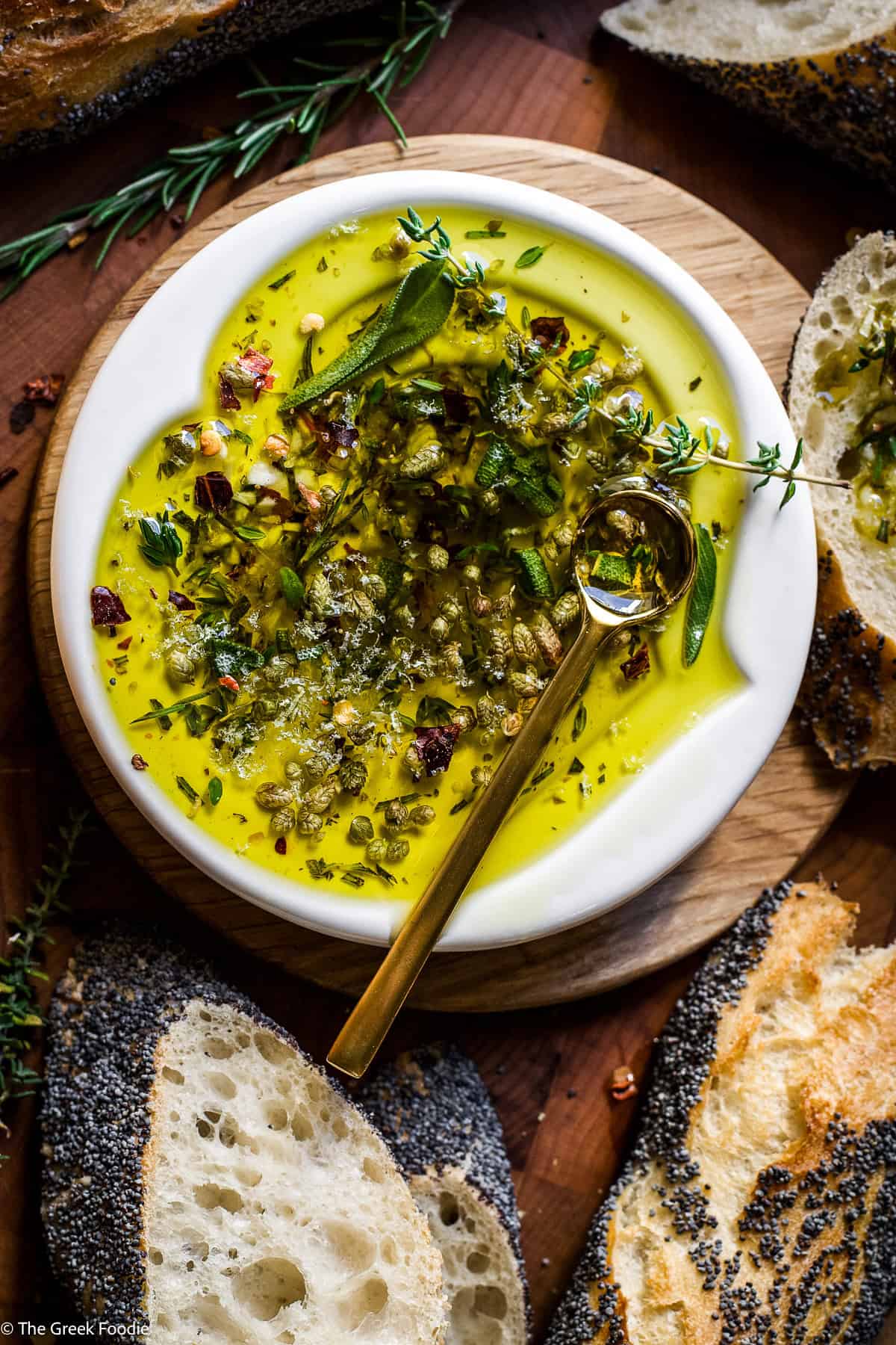 Overhead view of bread dipping oil with herbs and garlic in a small white dish, served with slices of poppy seed bread.