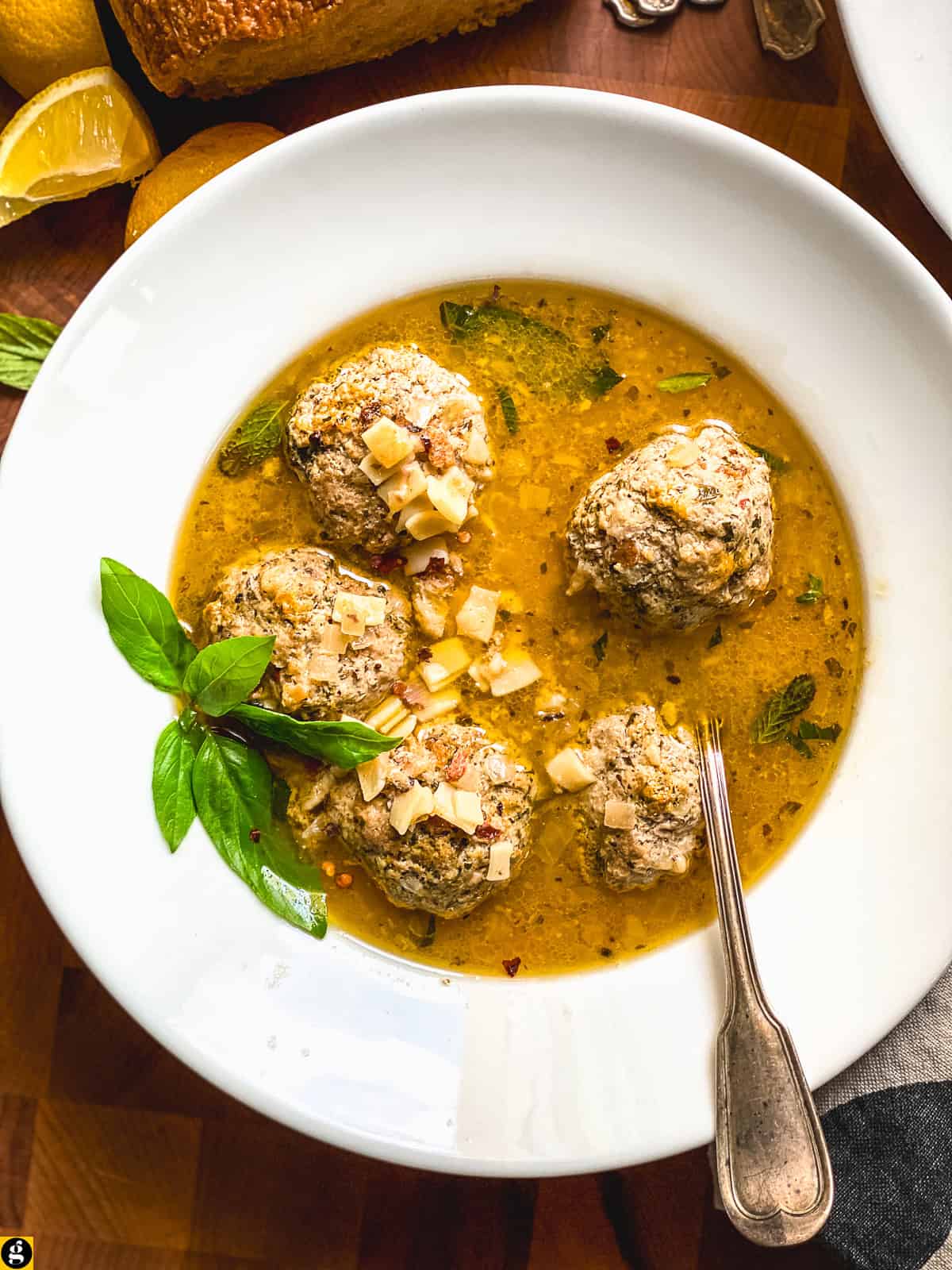 Overhead view of meatball soup with tiny square pasta, basil, mint, and chili flakes in a lemony broth, with lemons and bread nearby.