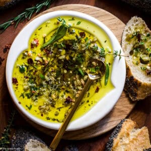 Olive oil bread dip with fresh rosemary, thyme, sage, and red pepper flakes in a white bowl with a gold spoon, surrounded by crusty bread.