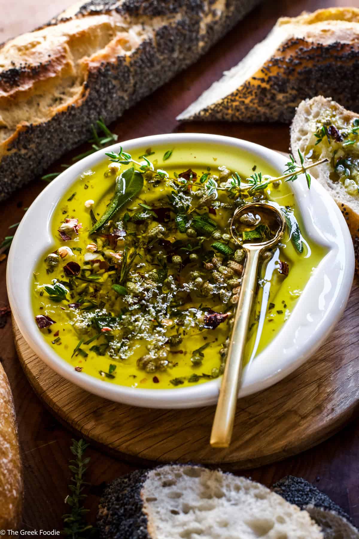 Close-up of olive oil dipping sauce for bread with fresh herbs, red pepper flakes, and a gold spoon in a white bowl.